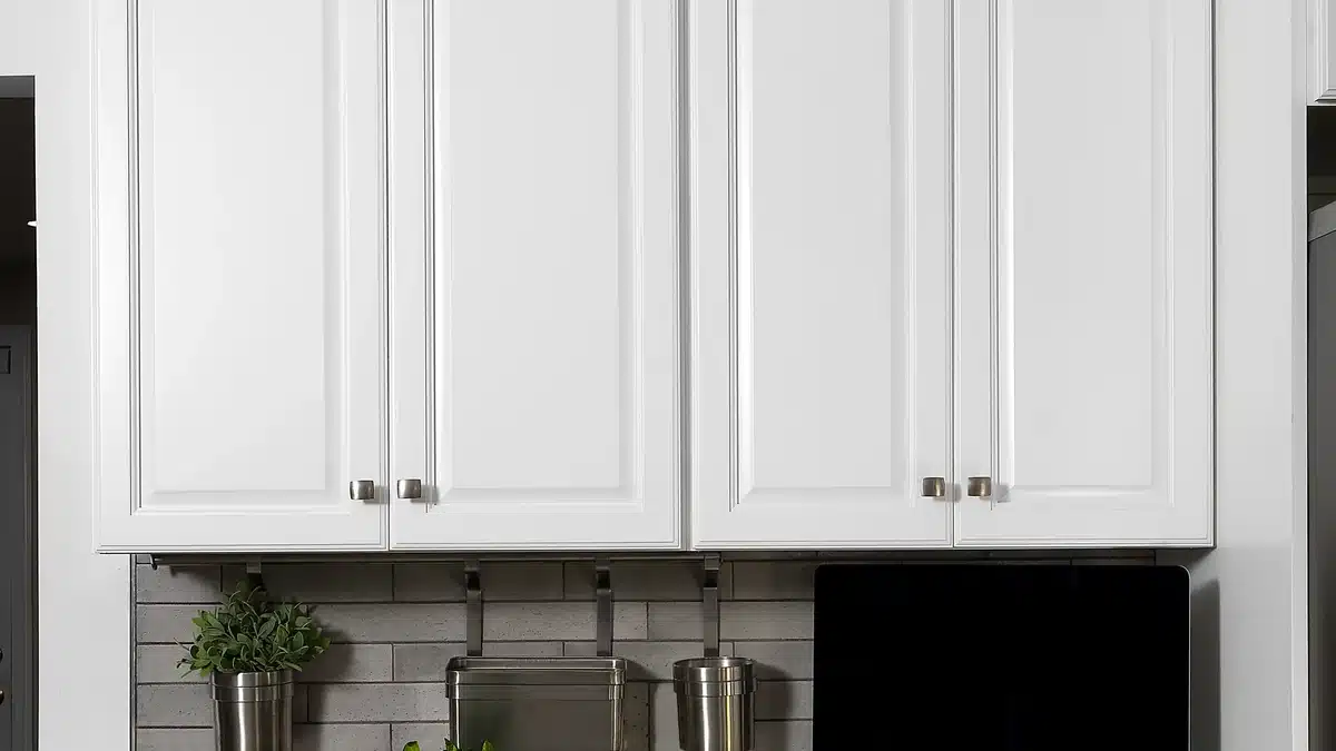 Close view of four white raised panel wall cabinet doors with small brushed metal knobs, hung above a grey brick style tile backsplash with a stainless steel rail holding utensil pots and a small potted plant.