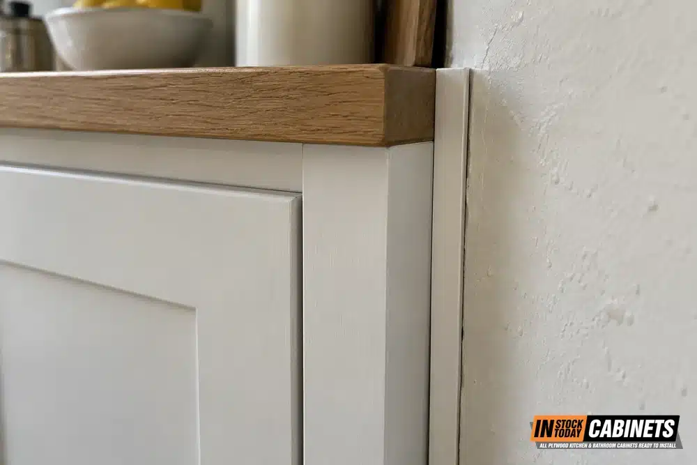 Close-up of the end of a run of white shaker kitchen cabinets under a wood countertop edge, where a narrow vertical strip in the same white finish closes the gap between the cabinet and a rough, unevenly painted plaster wall.